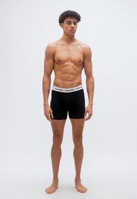 Muskelöser junger Mann steht barfuß da und trägt schwarze Boxershorts mit weißem Bund, auf dem "ONLY&SONS" steht, vor einem schlichten weißen Hintergrund.