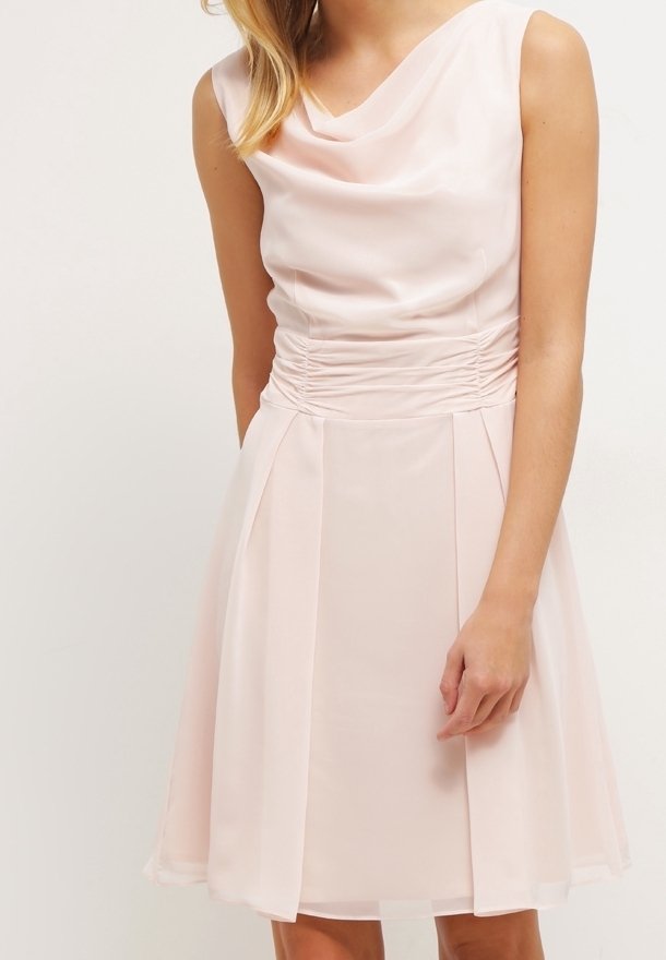 Robe rose pâle sans manches avec un décolleté drapé et une taille froncée portée par une femme sur un fond clair uni.