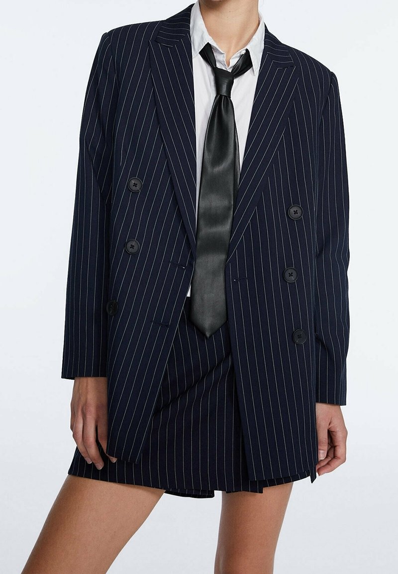 Personne portant un blazer croisé à rayures bleu marine avec une jupe assortie, une chemise blanche et une cravate noire, montré du cou à la mi-cuisse.