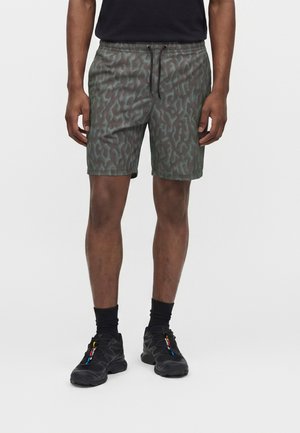 Mężczyzna noszący czarne buty sportowe, czarne skarpetki, zielone spodenki w wzory z sznurkiem oraz czarną koszulkę, stojący na jednolitym białym tle.