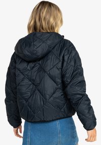 Veste matelassée noire à capuche, avec des motifs en losange et des poignets élastiques. Le dos est composé d'un matériau lisse et brillant.