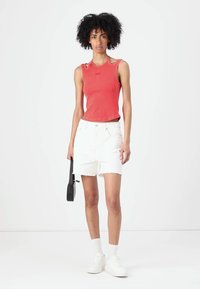 Rotes, geripptes Tanktop mit ausgeschnittenen Schulterdetails, kombiniert mit weißen Jeansshorts. Das Model hält eine schwarze Tasche und trägt weiße Sneaker und Socken.
