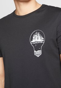 Černé bavlněné tričko s bílým potiskem designu lodi uvnitř grafiky žárovky na přední straně. Krátké rukávy, kulatý výstřih.