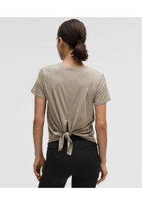 Beige short-sleeve top with a tied back design, featuring smooth fabric and a subtle logo near the hem. Paired with black leggings.