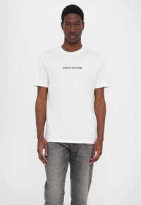 Valge lühikeste varrukatega T-särk, millel on must "ARMANI EXCHANGE" logo keskel rinnal. Kanda hallide teksadega. Lihtne, figuuri järgiv disain.