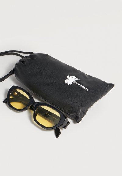 Lunettes de soleil à monture noire avec verres jaunes placées à côté d'une pochette noire à cordon présentant un logo de palmier blanc et le texte « palm angels ».