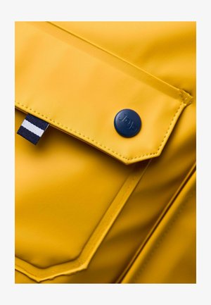 Tejido impermeable amarillo con un bolsillo frontal, con un botón azul marino etiquetado como "Jojo" y una etiqueta a rayas azul y blanca. Textura suave.