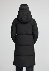 Svart puffjacka med huva, lång längd och quiltat mönster med horisontella sömmar. Slät textur, funktionell för kallt väder.