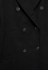 Crni dvostruki kaput s herringbone teksturom, četiri crna dugmeta i bočne džepove. Prilagođen kroj za strukturirani izgled.