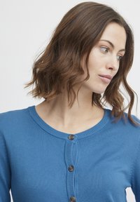 Cardigan tricotat albastru cu guler rotund, având butoane maronii pe față. Materialul pare moale, cu accente cu nervuri și o formă ajustată.