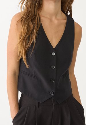 Mujer con chaleco negro sin mangas abotonado sobre pantalones negros a juego, con pelo largo, ondulado y castaño sobre un hombro.