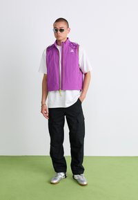 Man wearing purple sleeveless jacket, white T-shirt, black cargo pants, silver sneakers, and sunglasses standing on green floor against white wall.