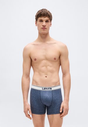 Jonge man staand, naar voren gericht, zonder shirt, draagt blauwe Levi's boxer briefs met witte tailleband tegen een effen lichte achtergrond.