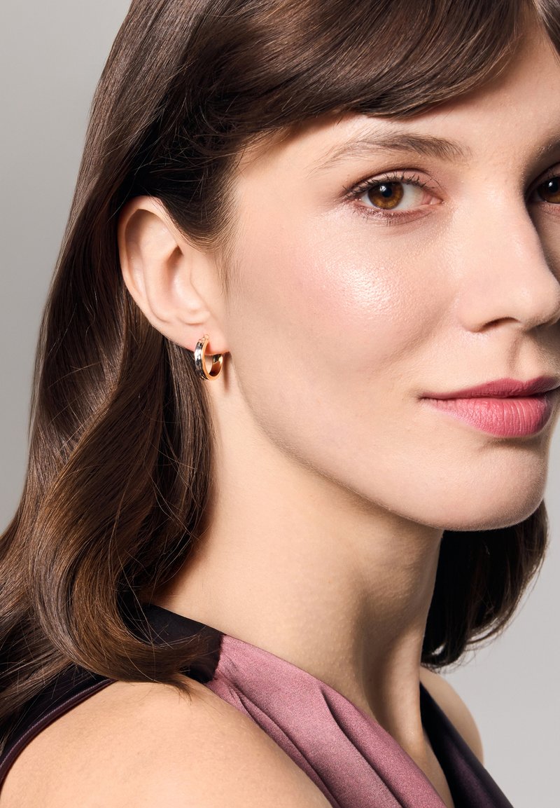 Złote kolczyki w kształcie kółek o gładkiej powierzchni, z małym srebrnym akcentem. Modelka z średniej długości brązowymi włosami, nosząca różowo-czarną odzież.