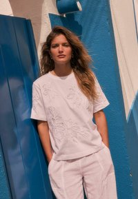 Un t-shirt blanc brodé avec des motifs floraux, assorti à un pantalon en lin de couleur claire. L'arrière-plan comprend un mur et une porte peints en bleu.