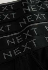 Fekete pamut alsónemű vastag derékpánttal, amelyen szürke betűkkel a "NEXT" felirat található. Az anyag puhanak tűnik, sima textúrával.