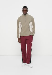 Lys beige halv-zip jakke med hvide sidepaneler, parret med bordeaux trackpants med sorte sidestriber og et logo.