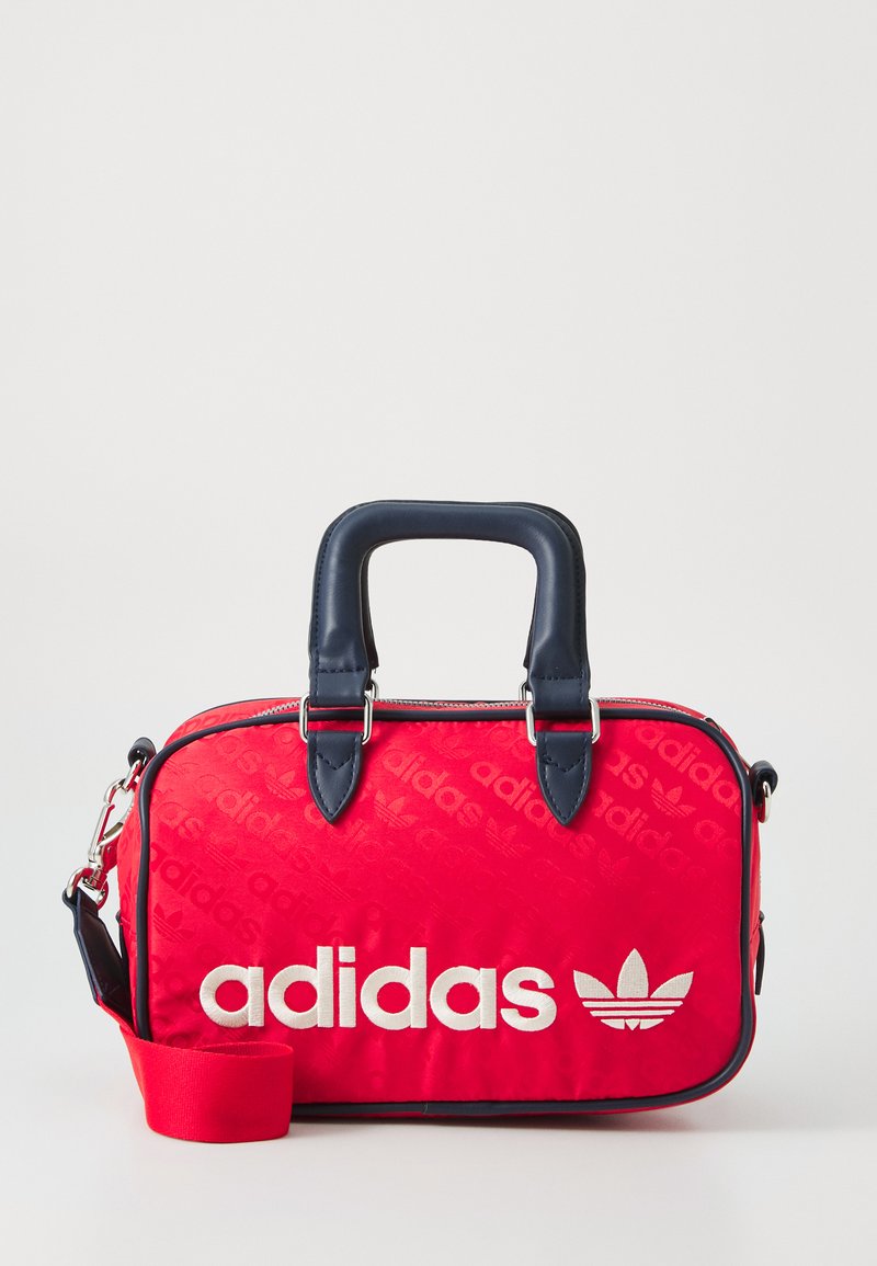 Czerwona torba z tkaniny z białym logo "adidas" i wzorem Trójlistnej Koniczyny, granatowe uchwyty oraz regulowany pasek. Zamek błyskawiczny u góry.