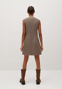 Robe sans manches à carreaux marron avec une silhouette ajustée, présentant un col montant et des détails de couture au dos. Complétée par des bottes marron montant jusqu'aux genoux.