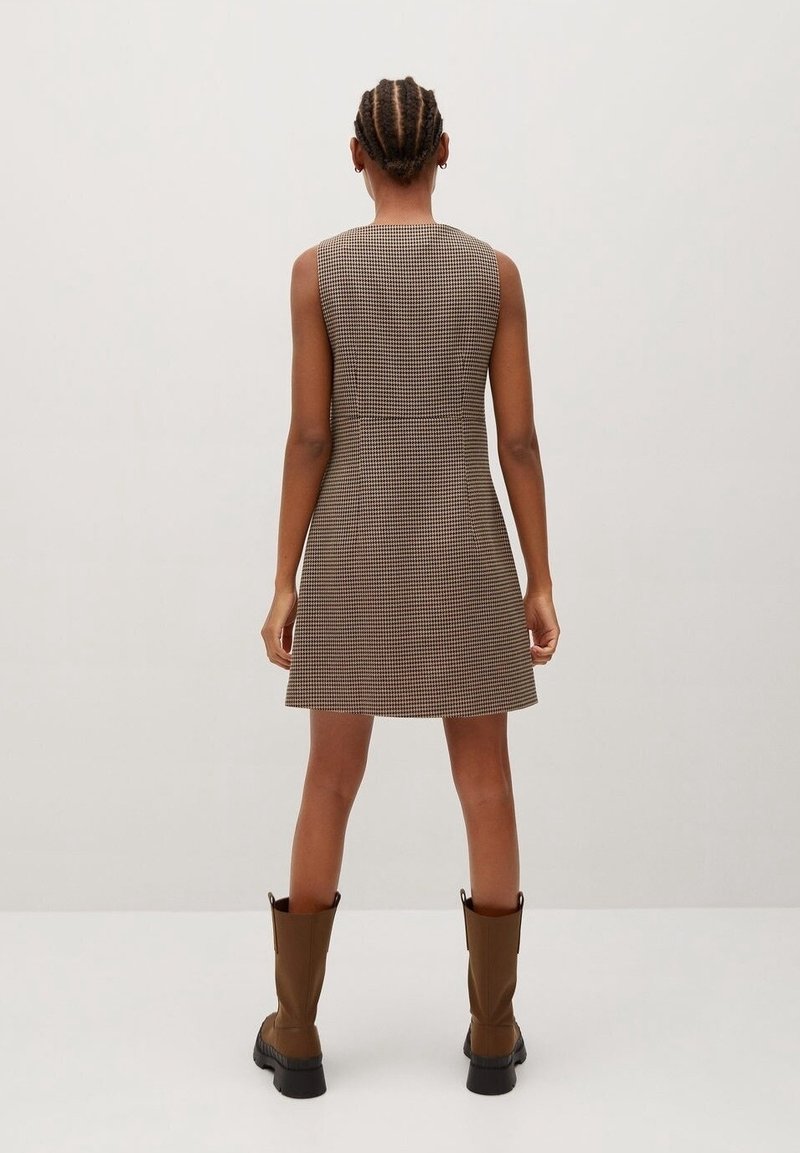 Robe sans manches à carreaux marron avec une silhouette ajustée, présentant un col montant et des détails de couture au dos. Complétée par des bottes marron montant jusqu'aux genoux.