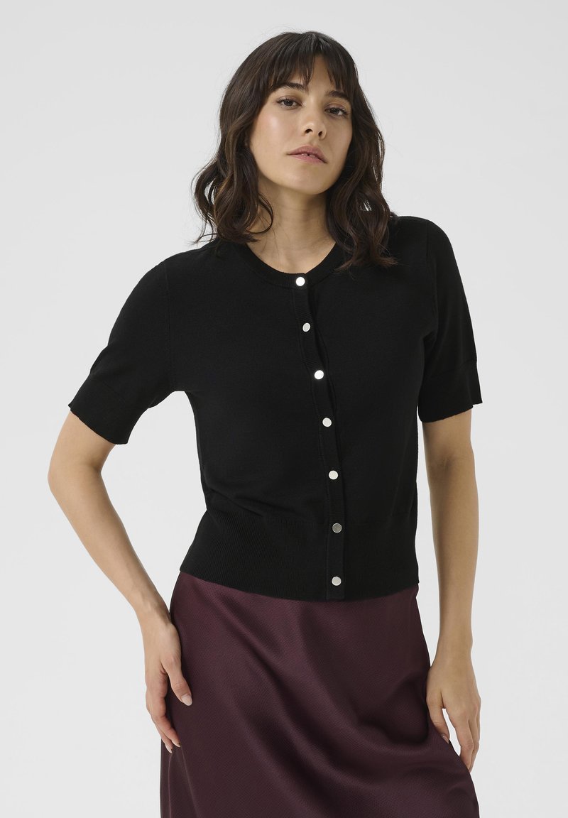 Black short-sleeve cardigan made of soft material, featuring a round neckline and silver-button front closure. Worn over a dark skirt.