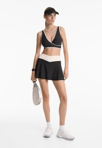 Crni sportski crop top s bijelim obrubom, u kombinaciji s crnom suknjom koja ima bež pojas i chevron uzorak; drži reketa.