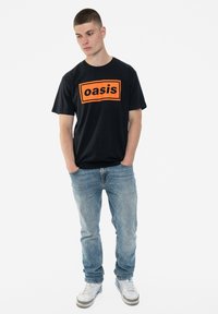 Black cotton t-shirt featuring an orange "oasis" logo in a rectangular design, paired with light blue jeans and white sneakers.