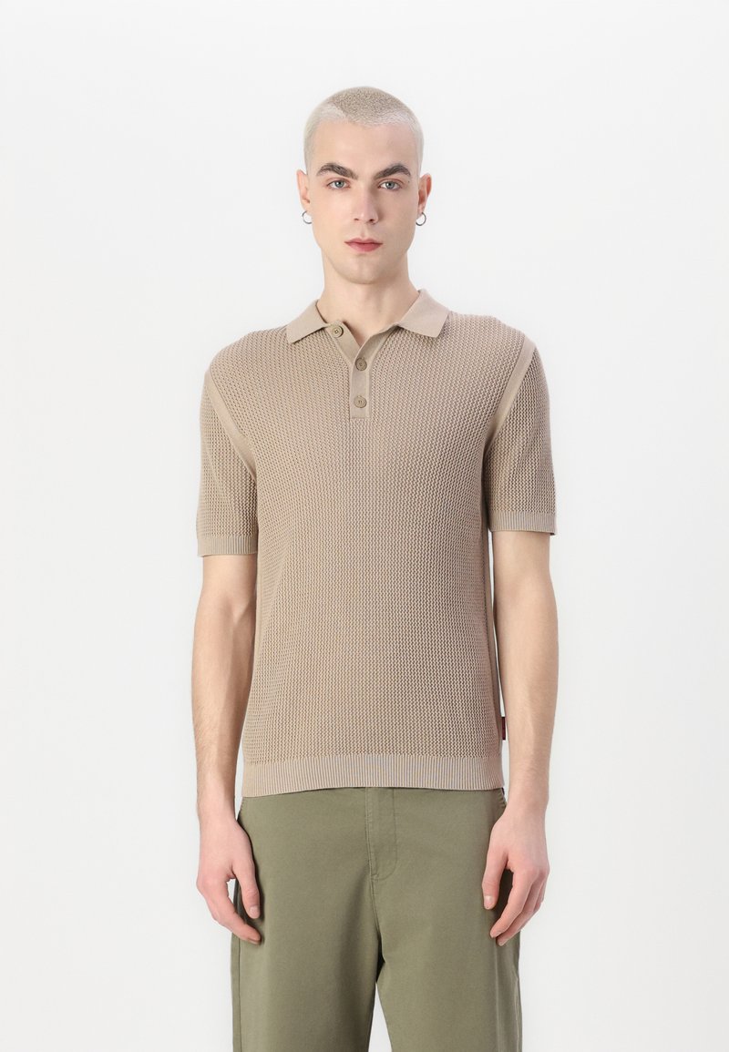 Beige polo s krátkým rukávem s texturovaným pleteným vzorem, přírodní tříknoflíkovou légou a žebrovanými detaily. Nosený s olivově zelenými kalhotami.