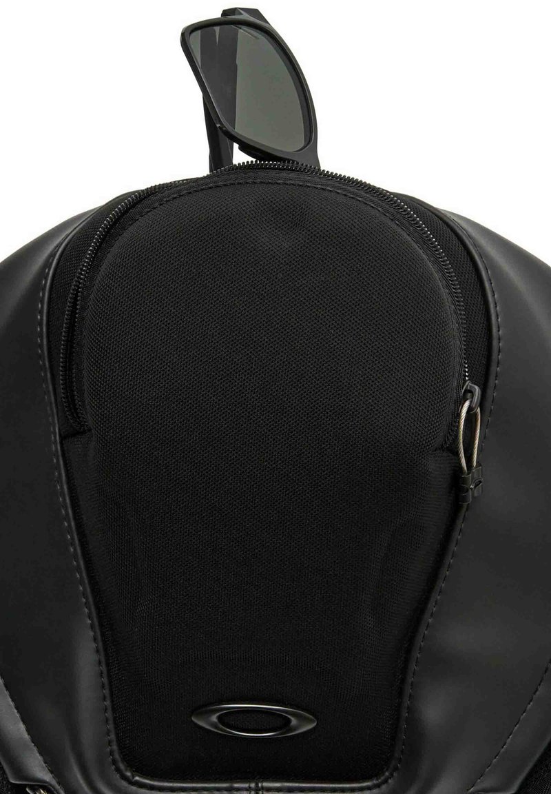 Zaino nero con una tasca con zip, fronte in tessuto testurizzato e dettagli in pelle liscia. Include occhiali da sole neri appoggiati sopra.