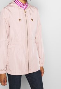 Ljusrosa zip-up jacka med huva, guldhardware detaljer, elastisk midja och samlade ärmar. Buren över en rosa och vit randig skjorta.