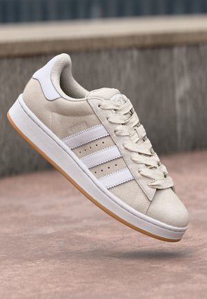 Baskets en daim beige et blanc avec des rayures blanches, semelle blanche épaisse avec semelle extérieure en gomme, lacets attachés, montrées flottant au-dessus d'un sol en pierre.