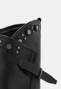 Zbliżenie na czarny skórzany but, z metalowymi srebrnymi ćwiekami, paskiem oraz lekko fakturowaną powierzchnią, podkreślające jego przemysłowe elementy designu.