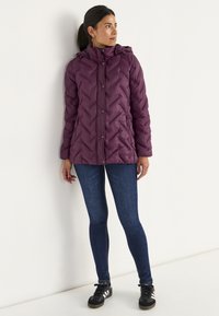 Burgundy puffjacka med huva, med chevron-quilting, tryckknappar och sidofickor; kombinerad med mörkblå slim fit jeans och svarta sneakers.