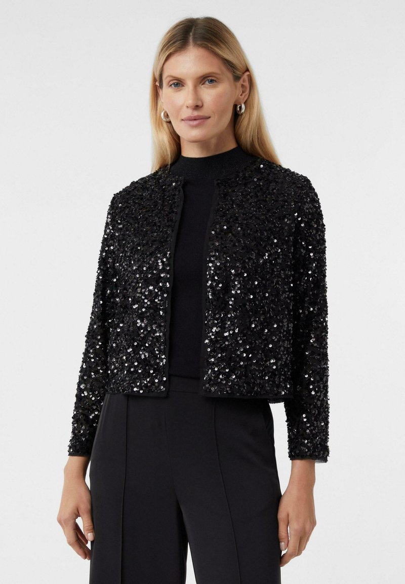 Kobieta w czarnej, błyszczącej kurtce z otwartym przodem, czarnym topie i czarnych spodniach, stojąca na tle białego tła.