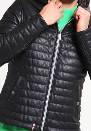 Persona con una chaqueta acolchada negra con cremallera y bolsillos con broches, ajustándose el cuello sobre una camisa verde.