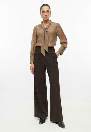 Woman stands wearing brown textured blouse with necktie detail, dark wide-leg trousers, and black pointed shoes on plain white background.