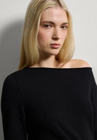 Maglione nero off-the-shoulder con maniche lunghe, realizzato in morbido tessuto a maglia. Texture liscia e design minimalista con una vestibilità rilassata.