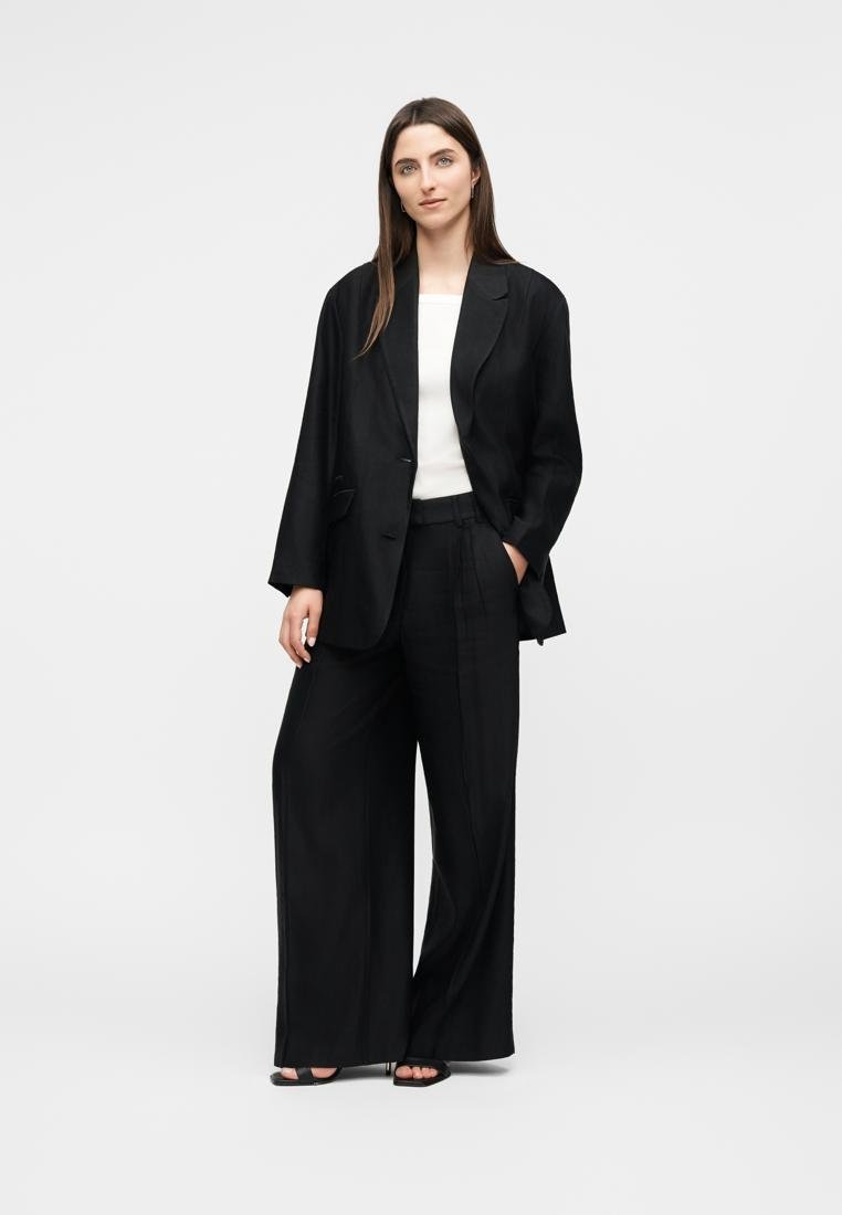 Femme debout, portant un blazer noir ample, un pantalon noir à jambes larges, un haut blanc et des talons ouverts noirs, sur un fond clair uni.