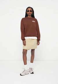 Sweatshirt castanho com texto branco, sobre uma camisa branca. Saia beige, meias brancas e sapatilhas cinzentas completam o look. Óculos de sol pretos usados.