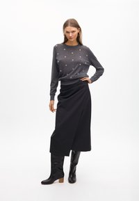 Grauer Pullover mit Perlendetails, kombiniert mit einem langen schwarzen Rock. Das Modell trägt kniehohe schwarze Stiefel mit einem quadratischen Absatz. Schlichter Hintergrund.