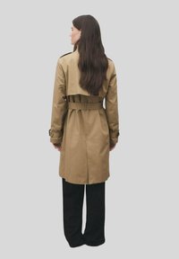Cappotto beige con vita aderente, piega sul retro e cintura. Maniche lunghe con polsini regolabili. Indossato sopra pantaloni neri.