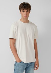 Lichtbeige katoenen t-shirt met korte mouwen, ronde hals en subtiele logo op de borst; gecombineerd met blauwe jeans. Glad textuur.