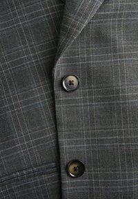 Sivo karirano blago jakne s dvema črnima gumboma in vidnim detajlom majhnega žepa.