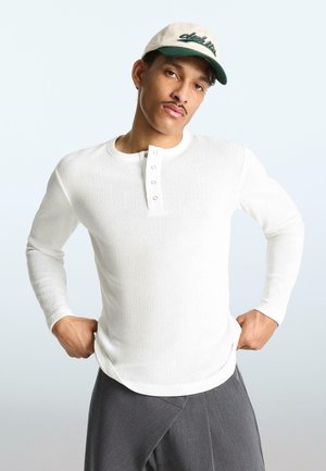 Fehér hosszú ujjú henley-inget, szürke nadrágot és bézs sapkát viselő férfi, kezét a csípőjén tartva, egyszínű világos háttér előtt áll.