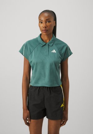 Türkises Polo-Shirt mit kurzen Ärmeln und Kragen, das ein strukturiertes Muster und ein weißes Logo aufweist. Kombiniert mit schwarzen Shorts und gelben Akzenten.