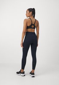 Soutien de desporto preto com costas recortadas, combinado com leggings azul-marinho de cintura alta com acabamentos texturizados. Modelo usa sapatilhas de desporto pretas.