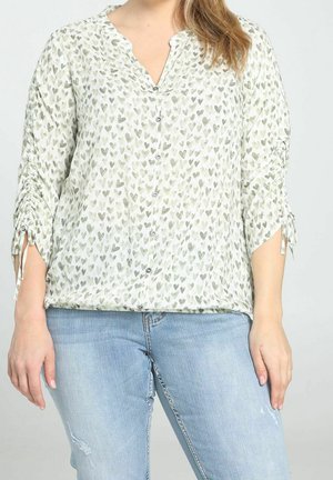 Femme portant un chemisier blanc à boutons avec des motifs de cœurs gris et des manches froncées, associé à un jean bleu clair usé.