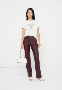 Camiseta blanca con adornos en forma de triángulo plateado, pantalones de cuero sintético burdeos, bolso blanco y sandalias de tacón beige con acentos de logo.