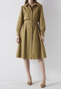 Mujer vistiendo un vestido midi verde olivo de manga larga con cinturón y botones en la parte delantera, combinándolo con tacones abiertos verdes a juego, de pie sobre un fondo blanco.