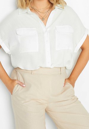 Kobieta ubrana w białą, krótką koszulę zapinaną na guziki z dwoma kieszeniami na piersi oraz beżowe, wysokie spodnie, ręce w kieszeniach.